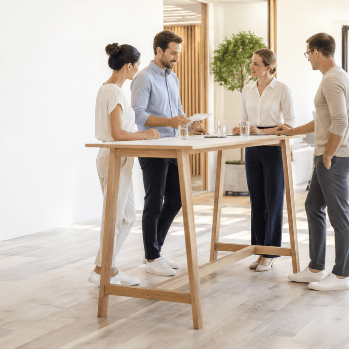 Vier Kolleginnen und Kollegen bei einem kurzen Stand-up-Meeting an einem hellen Holz-Stehtisch; eine Person zeigt ein Blatt Papier, während durch geöffnete Fenster warmes Sonnenlicht in das moderne Büro fällt.